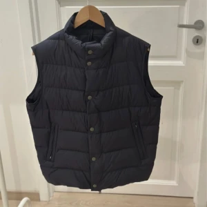 Blå dunväst från Massimo Dutti - Stilren svart dunväst från Massimo Dutti med hög krage och tryckknappar framtill. Västen har två dragkedjeförsedda fickor och quiltad design som ger en modern look. Perfekt för lager-på-lager under kyliga dagar.