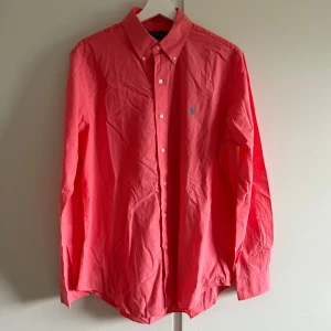 Rosa skjorta från Polo Ralph Lauren - Klassisk rosa skjorta från Polo Ralph Lauren med button down-krage och blå broderad logga på bröstet. Skjortan har långa ärmar, knäppning framtill och är tillverkad i mjuk bomull. Perfekt för en stilren och fräsch look.