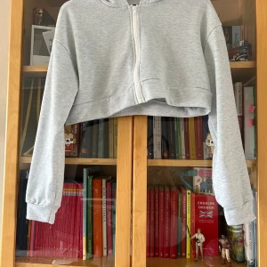Grå croppad hoodie med dragkedja - Snygg ljusgrå croppad hoodie med dragkedja framtill och huva. Tröjan har långa ärmar med breda muddar och är tillverkad i mjukt sweatshirtmaterial. Perfekt för en avslappnad och trendig look.