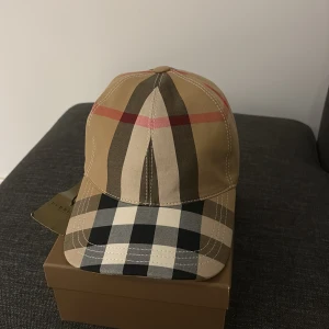 Burberry rutig keps i beige och svart - Snygg och unik keps från Burberry med klassiskt rutigt mönster i beige, svart, vitt och röda detaljer. Kepsen har böjd skärm och justerbar baksida. Tillverkad i bomull och perfekt för dig som vill ha en stilren accessoar med lyxig känsla. PRIS KAN DISKUTERAS 