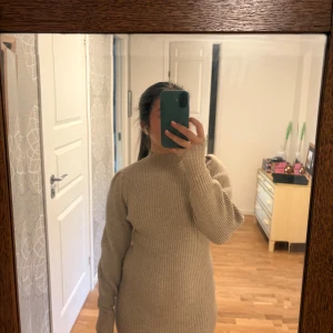 Beige ribbad stickad klänning - Supermysig beige stickad klänning med ribbat mönster och höghalsad krage. Modellen är kort och har långa ärmar med lätt puff vid axlarna. Snygg att styla med boots eller sneakers.