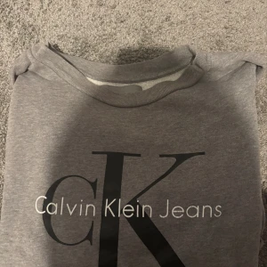 Grå Calvin Klein Jeans sweatshirt S - Snygg grå sweatshirt från Calvin Klein Jeans i storlek S. Tröjan har ett stort CK-tryck i svart och vitt framtill och är tillverkad i mjuk bomullsmix. Rund halsringning och långa ärmar gör den perfekt för chill dagar. Enkel och stilren design som passar till jeans eller joggers.