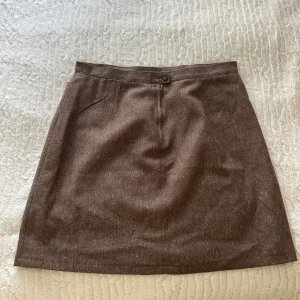 Brun ullkjol från Sinclair by Kappahl - Snygg brun kortkjol från Sinclair by Kappahl i klassiskt snitt. Kjolen är tillverkad i ett mjukt ullblandat tyg med diskret fiskbensmönster och har en enkel knapp- och dragkedjestängning framtill. Perfekt för dig som gillar stilrena och tidlösa plagg.