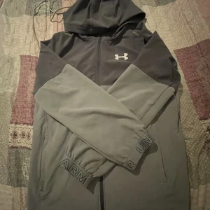 Grå vindjacka från Under Armour - Snygg grå vindjacka från Under Armour med huva och hel dragkedja framtill. Jackan har två sidofickor, elastiska muddar, jackan passar den som är mellan 146,152 cm lång antingen så kan du köpa den för sig själv eller ge den till Din släkting 