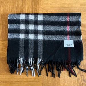 Rutig halsduk från Burberry svart  - Snygg halsduk från Burberry i klassiskt rutigt mönster med svarta, gråa och röda toner. Halsduken har fransar i ändarna och är tillverkad i mjuk ull. Perfekt accessoar för att lyfta din outfit.