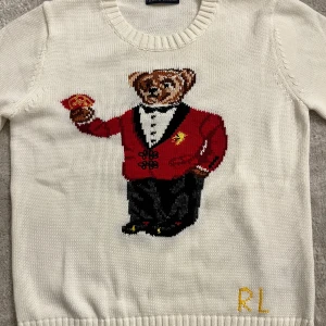 Vit stickad tröja med björn från Polo Ralph Lauren - Vit stickad tröja från Polo Ralph Lauren med ikonisk björn i röd kavaj broderad på framsidan. Rund halsringning och ribbade muddar. RL-broderi i gult vid nederkanten.