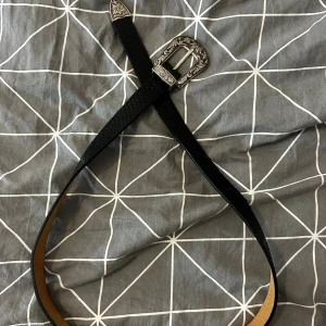 Svart skärp med silverdetaljer - Snyggt svart bälte i skinn med reptilmönstrad yta och coolt silverfärgat spänne med graverade detaljer. Bältet har en smal form och spetsig avslutning, perfekt för att ge din outfit en extra edge. Passar dig som gillar western vibes och detaljer.