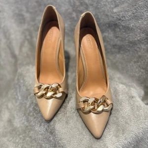 Beige pumps med guldkedja från Kazar - Snygga beige pumps från Kazar med spetsig tå och hög klack. Skorna är i skinn och har en chunky guldfärgad kedjedetalj framtill som ger en lyxig vibe. Perfekta för dig som vill sticka ut med stilrena men ändå trendiga klackar.