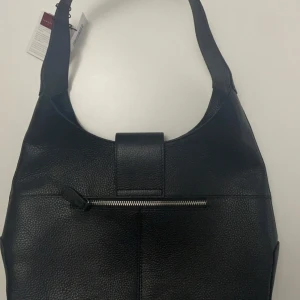 Svart axelväska från Lamarthe i skinn - Stilren svart axelväska från Lamarthe i äkta skinn med silverfärgade metalldetaljer och dragkedja framtill. Väskan har ett rymligt foder med logomönster och flera innerfack med dragkedja. Perfekt för dig som vill ha en klassisk och praktisk väska.