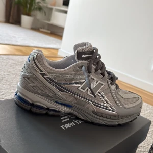 New Balance 1906 grå sneakers - Säljer ett par New Balance 1906 sneakers i grått med silvriga detaljer och meshpaneler. Skorna har snörning, platt sula och en sportig vibe. Perfekta för dig som gillar stilrena och bekväma sneakers med retroinspirerad design. Helt nya 10/10 skick från JD sports 