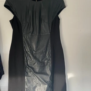 Svart klänning med skinnpanel - Snygg svart klänning med cool skinnpanel framtill och på axlarna. Klänningen har en rak passform och korta ärmar. Klänningen är i bra skick men skinnet har blivit lite skrynkligt som man ser på bilden! 