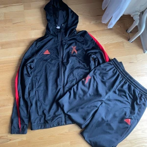 Svart Adidas hoodie med röda detaljer - Svart Adidas hoodie med huva och röda detaljer längs ärmarna samt stora X-tryck på bröstet och ryggen. Tröjan har dragkedja framtill och är tillverkad i mjuk polyester, perfekt för träning eller chill. Matchande svarta byxor med röda detaljer och Adidas-logga ingår.