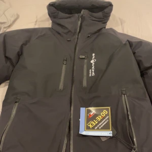 Svart Gore-Tex vindjacka Sail Racing - Svart vindjacka från Sail Racing med Gore-Tex-material, perfekt för blåsiga dagar. Jackan har flera dragkedjeförsedda fickor, hög krage och justerbar huva. Snygg logga på bröstet och stilren design som passar till streetwear.