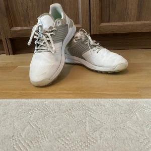 Vita golfskor från Puma - Säljer ett par vita golfskor från Puma med grå detaljer och snörning. Skorna har en sportig design med rund tå och platt sula, samt blå dobbar undertill för extra grepp. Tillverkade i syntetmaterial och mesh för bra andningsförmåga.