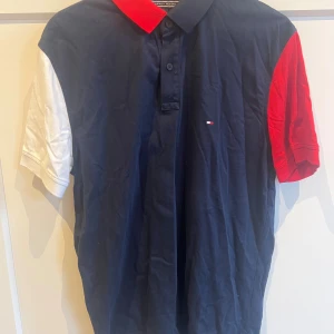 Tommy Hilfiger färgblockad piké L - Snygg pikétröja från Tommy Hilfiger i storlek L. Tröjan har marinblå bas, röd krage, ena ärmen i rött och den andra i vitt. Klassisk knappslå framtill och liten broderad logga på bröstet. Regular fit och tillverkad i bomull.