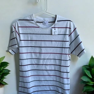 Vit randig t-shirt från Bläck  - Snygg vit t-shirt från Bläck med tunna horisontella ränder i svart, grått och vinrött. Klassisk rund hals och korta ärmar. Tillverkad i mjuk bomull som känns skön mot huden. Perfekt för dig som gillar enkel och stilren design.