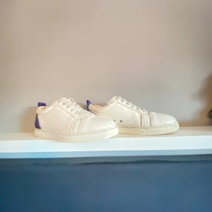Vita Louboutin sneakers med lila detalj - Säljer ett par vita sneakers från Christian Louboutin med klassisk röd sula och lila detalj på hälen. Skorna har snörning, rund tå och är tillverkade i skinn. Perfekta för dig som vill sticka ut med en lyxig touch och ikonisk design.