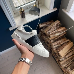 Valentino Garavani  - Snygga Valentino Garavani Open sneakers i vitt skinn med bred svart läderdetalj över mitten. Klassisk rund tå, platt sula och coola nitar baktill på hälen. Skorna har snörning och en stilren, lyxig vibe som passar perfekt till streetwear eller mer dressade outfits.
