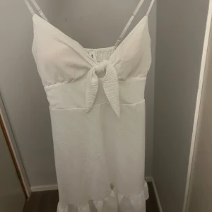Vit sommarklänning med knyt framtill - Superfin vit klänning från H&M med smala axelband och knyt framtill. Klänningen har en lätt rynkad struktur och volang nertill, perfekt för varma dagar. Enkel och stilren design som känns både fräsch och trendig.