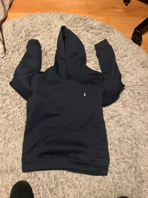 Mörkblå hoodie från Polo Ralph Lauren - Snygg mörkblå hoodie från Polo Ralph Lauren med klassisk broderad logga på bröstet. Tröjan har huva med snörning, stor magficka och långa ärmar. Perfekt för chill dagar och enkel att matcha med jeans eller joggers.