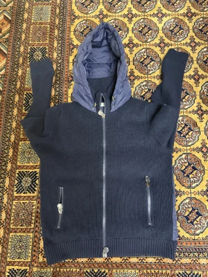 Moncler cardigan  - Snygg mörkblå jacka från Moncler med stickade ärmar och framsida, samt quiltad rygg och huva i glansig polyester. Jackan har dragkedja framtill och två fickor med dragkedja. Perfekt för dig som vill ha en sportig och exklusiv look.