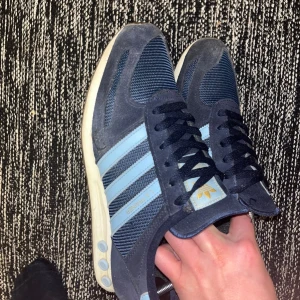 Adidas LA Trainer sneakers marinblå - Säljer ett par Adidas LA Trainer sneakers i marinblått med ljusblå ränder och detaljer. Ovandelen är i mesh och mocka, med klassisk snörning och vit sula med de ikoniska pluggarna på sidan. Perfekta för dig som gillar retrostil och sportig vibe. (KOM MED BUD!)
