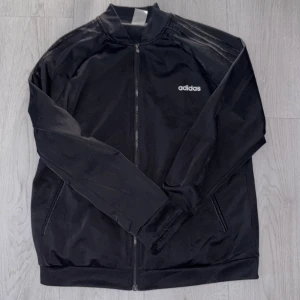 Svart Adidas zip-tröja XL - Svart zip-tröja från Adidas i storlek XL med klassiska tre ränder på axlarna och vit Adidas-logga på bröstet. Tröjan har lång ärm, ståkrage och dragkedja hela vägen. Materialet är mjukt och lätt polyester, perfekt för träning eller chill.