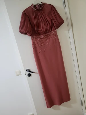 Röd långklänning med pärlor - Säljer en elegant röd långklänning med puffiga korta ärmar och hög hals dekorerad med pärlor. Klänningen har plisserad överdel och pärldetaljer som fortsätter ner över midjan. Perfekt för dig som vill sticka ut med en unik och lyxig look.