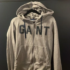 Grå hoodie från GANT med dragkedja - Säljer en grå hoodie från GANT i storlek S med blå snören och snygg grisch logga framtill. Tröjan har huva, fickor och dragkedja hela vägen. Har en klassisk grisch stil. 