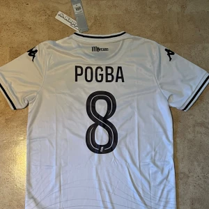 AS Monaco Pogba #8 vit fotbollströja - Snygg vit AS Monaco fotbollströja från Kappa med Pogba och nummer 8 på ryggen. Tröjan har svarta detaljer vid krage och ärmslut, klubbmärke på bröstet och sponsortryck. Tillverkad i lätt och ventilerande polyester, perfekt för match eller träning.