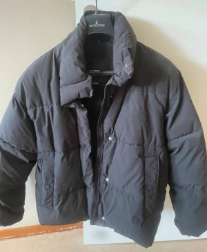 Svart pufferjacka från Pull&Bear - Svart pufferjacka från Pull&Bear med hög krage och quiltad design. Jackan har dragkedja och tryckknappar framtill, samt två sidofickor. Materialet är syntet och jackan har en loose passform som ger en chill vibe. Perfekt för kalla dagar.