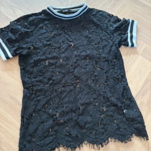 Svart spetsblus från Bikbok, S - Svart blus från Bikbok i storlek S, gjord i genombruten spets med blommigt mönster. Ribbad krage och ärmslut i vitt och svart ger en sportig touch. Kortärmad och med snygg scallop-kant nedtill. Perfekt för dig som gillar att sticka ut.