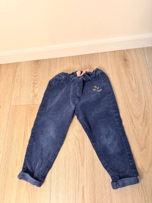 Blå manchesterbyxor för barn - Mörkblå manchesterbyxor från 5.10.15 i storlek 80. Byxorna har en rosa snörning i midjan och söta broderade blommor i olika färger på ena benet. Modellen är loose fit med uppvikta benslut och mjuk, skön känsla.