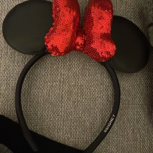 Disney diadem med svarta öron och röd paljettrosett har aldrig haft på mig dem köpte dem till Halloween men jag hittade en bättre kostym - Säljer ett ikoniskt Disney diadem med stora svarta musöron och en glittrande röd rosett täckt av paljetter. Perfekt för dig som vill sticka ut på temadagar eller bara vill ha något extra till din look. Diademet är bekvämt och sitter snyggt på huvudet.