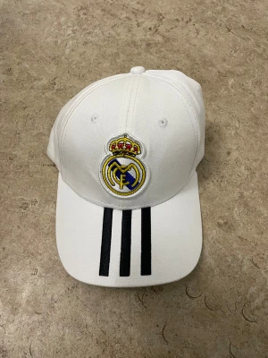 Vit Real Madrid keps från Adidas - Snygg vit keps från Adidas med Real Madrid-emblem broderat framtill och tre svarta ränder på skärmen. Justerbar baktill för perfekt passform. Perfekt för dig som älskar fotboll och vill representera ditt favoritlag med stil.