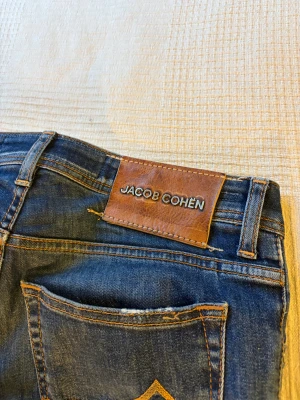 Blå jeans från Jacob Cohen - Snygga blå jeans från Jacob Cohen med klassisk femficksdesign och slitna detaljer. Jeansen har orange kontrastsömmar och ett brunt läderpatch med logga bak. Modellen är rak och materialet är mjukt denim i bomull.