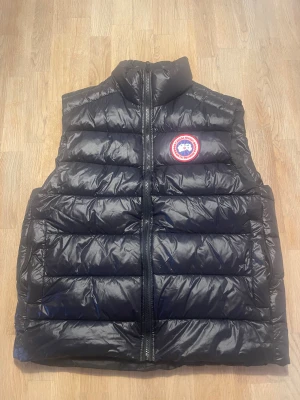 Canada Goose väst - Säljer en svart dunväst från Canada Goose med klassisk logga på bröstet. Västen har quiltad design, hög krage och dragkedja framtill. Perfekt för lager-på-lager och har en clean look med diskreta reflexdetaljer på ryggen. Grym att styla till hoodie eller långärmad.