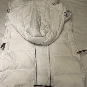 Vit pufferväst från Tommy Hilfiger - Säljer en vit pufferjacka från Tommy Hilfiger med huva och coola detaljer i rött, vitt och marinblått på muddar och fickor. Jackan har tvåvägsdragkedja och är tillverkad i polyester, perfekt för kalla dagar. Klassisk och sportig look med snygga Hilfiger-ränder.