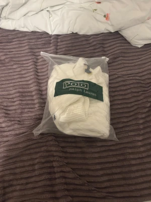 Vit kabelstickad tröja Polo Ralph Lauren - Vit kabelstickad tröja från Polo Ralph Lauren i storlek L. Klassisk design med rund hals och ribbade muddar vid ärmslut och nederkant. Perfekt för dig som gillar stilrena och tidlösa plagg med premiumkänsla.