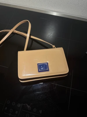 Beige axelväska från Acne Studios - Snygg beige axelväska från Acne Studios i slätt skinn med justerbar rem och magnetlås. Väskan har en stilren design med ett blått kvadratiskt emblem på framsidan och praktiska fack inuti, inklusive dragkedja. Perfekt för dig som gillar minimalistisk stil.