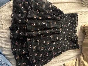 Svart blommig volangkjol Bik Bok XS - Supersöt svart kortkjol från Bik Bok med små blommor i rosa, vitt och gult. Kjolen har volanger och en smockad midja som sitter skönt och ger en snygg siluett. Perfekt för dig som gillar romantisk stil och vill ha något som sticker ut lite extra.