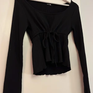 Svart långärmad blus med knyt - Svart blus med lång ärm och snygg knytning framtill. Toppen har en lätt rynkad struktur och en något croppad passform med vågig kant nedtill. Perfekt för dig som gillar stilrena och trendiga plagg med lite extra detalj.