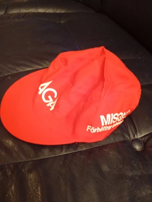 Röd AGA keps med tryck - Säljer en röd keps med vit AGA-logga och texten 'Förbättra arbetsmiljön' tryckt på sidan. Kepsen har klassisk form med böjd skärm och är tillverkad i lätt bomullstyg. Perfekt för dig som gillar retro eller vill sticka ut med en unik accessoar.