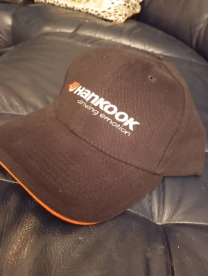 Svart Hankook keps med orange kant - Snygg svart keps från Hankook med broderad logga i vitt och orange framtill. Kepsen har en böjd skärm med en cool orange kant och klassisk form. Perfekt för dig som gillar motorsport eller vill ha en unik accessoar.