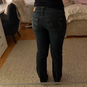 Mörkblå True Religion jeans straight - Säljer ett par mörkblå jeans från True Religion med klassiska kontrastsömmar och ikoniska bakfickor med hästsko-design. Modellen har rak passform och låg midja, med detaljerade nitar och knappstängning framtill. Perfekt för dig som gillar street och premium denim.