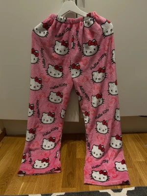 Hello Kitty rosa mjukisbyxor - Supermysiga rosa mjukisbyxor med Hello Kitty-tryck över hela byxan. Byxorna har elastisk midja och är gjorda i mjuk fleece som är perfekt för chill hemma. Söt design med vita Hello Kitty-ansikten och röda rosetter. (short)