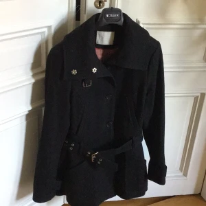 Svart kappa med bälte från BZR - Svart varm ,äkta wool kappa från BZR of Sweden med bred krage, dekorativa knappar och ett snyggt bälte med metallspänne i midjan. Jackan har långa ärmar, fickor framtill och klassisk, stilren design. Perfekt för dig som gillar en clean look. 