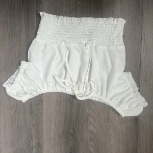 Vita shorts med smock och snörning - Säljer ett par vita shorts med bred smock i midjan och benöppningarna. De har en söt snörning framtill och är gjorda i ett mjukt, lätt tyg som känns luftigt. Passformen är loose och midjan är hög, perfekt för sommarens varma dagar.