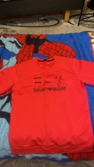 Röd sweatshirt från Emporio Armani - Säljer en röd långärmad sweatshirt från Emporio Armani med EA7-logga i svart och rött framtill. Tröjan har rund hals och är tillverkad i mjukt bomullsmaterial, perfekt för en chill och snygg look.