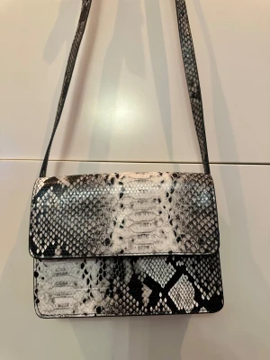 Axelväska med ormmönster från Pieces - Snygg axelväska med rektangulär form och magnetlås från Pieces. Väskan har ett svartvitt ormmönster och är tillverkad i syntetmaterial. Perfekt storlek för det viktigaste och har en lång axelrem. Stilren och trendig design som sticker ut.